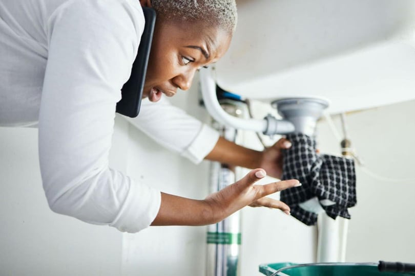 A woman on the phone looking worried as she holds rags against the pipes under the kitchen sink